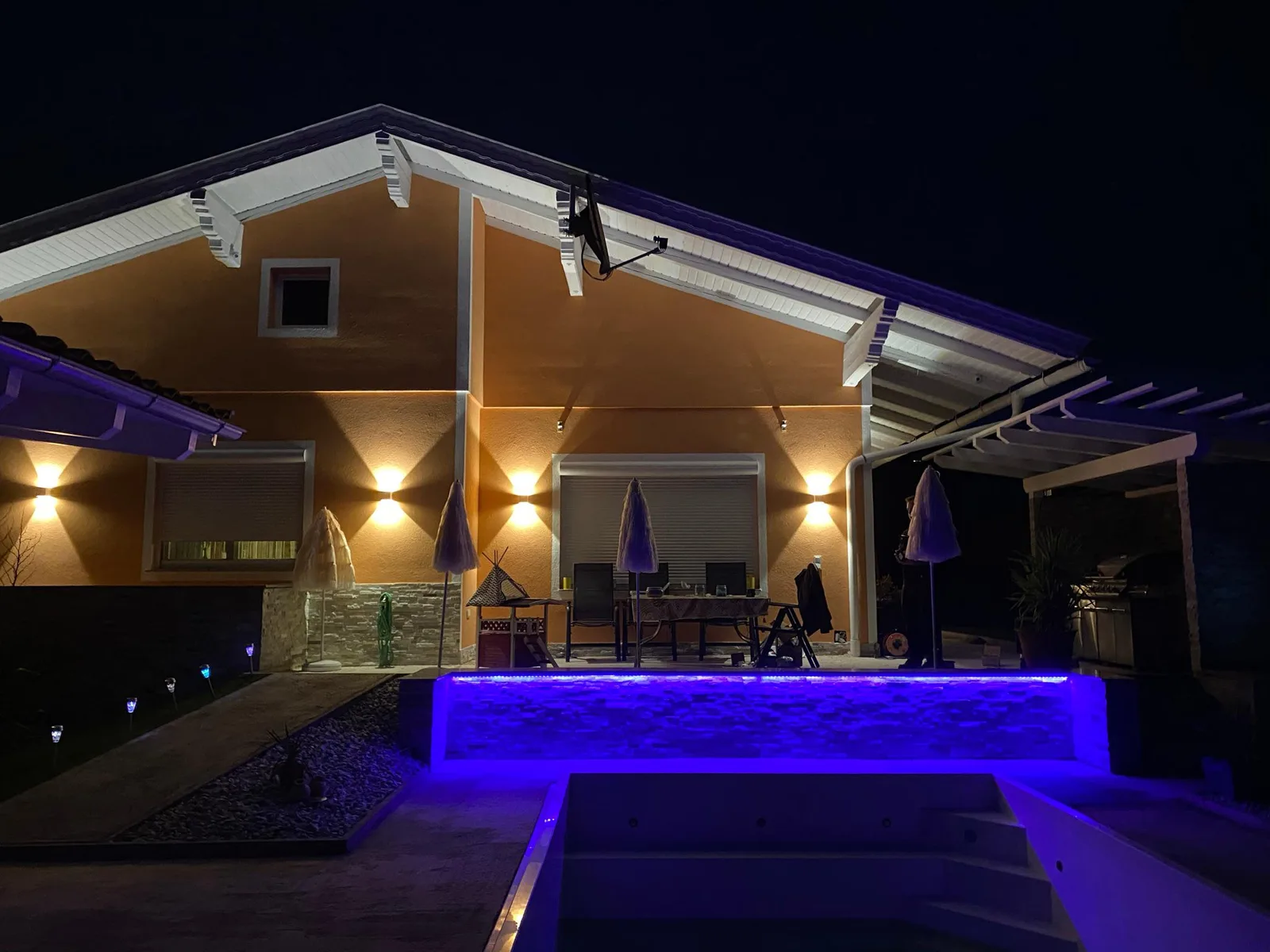 Wohnhaus bei Nacht mit warmen Wandleuchten an der Fassade und blau beleuchteter Mauer am Pool