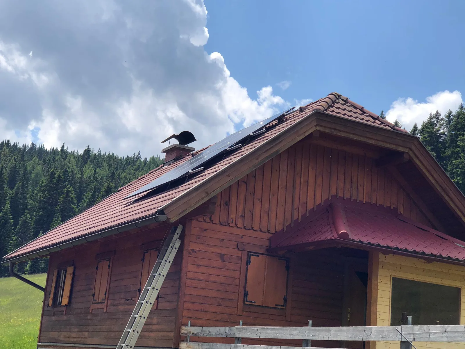 Holzhütte mit rotem Ziegeldach und Photovoltaikmodulen vor Nadelwald, eine Leiter an der Wand